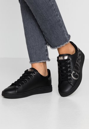Persona con zapatillas negras de caña baja con el logo "Guess" en los laterales y detalles metálicos, combinadas con vaqueros grises recortados con dobladillo deshilachado.