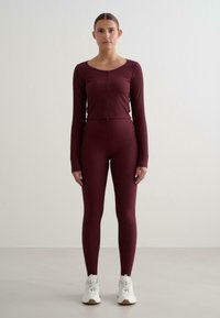 Bordeauxrotes Sportoutfit mit einem figurbetonten Langarmshirt mit Frontreißverschluss und hoch taillierten Leggings. Weiße Sneaker runden den Look ab.