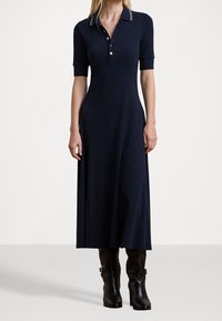 Mörkblå poloklänning med korta ärmar, sidöppningar, knäppt krage och kontrasterande kragslipsar, kombinerad med knähöga svarta stövlar.