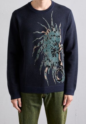 Mann trägt einen marineblauen Pullover mit großem türkis- und beigem abstraktem Blumenmuster und olivgrüne Hose, steht vor einer grauen Wand.