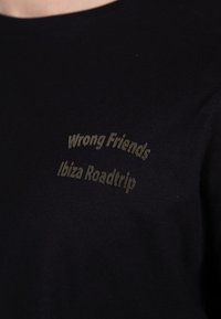 Sort bomulds T-shirt med guldtekst: "Forkerte Venner" og "Ibiza Roadtrip" i et buet design, med en glat tekstur.