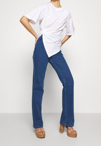 Persona vistiendo una camiseta blanca con nudos de manga corta, jeans de mezclilla azules y sandalias de plataforma de tacón alto beiges, de pie contra un fondo liso.