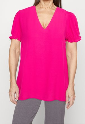 Vrouw die een felroze V-hals blouse met korte pofmouwen en grijze geribde broek draagt tegen een effen achtergrond.