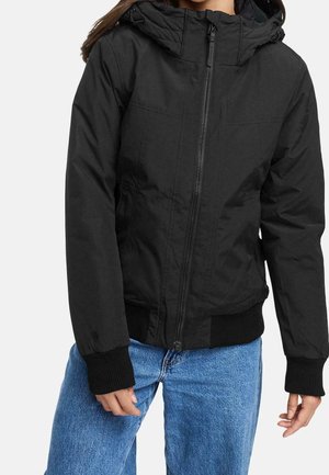 Frau trägt schwarzen Kapuzen-Reißverschlussjacke mit gerippten Bündchen und Saum, kombiniert mit blauen Jeans, steht vor einfachem Hintergrund.