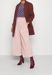 Manteau marron sur un haut bleu à motifs, associé à un pantalon large rose clair et des bottes à talons bourgogne. Textures lisses et coupe ajustée.