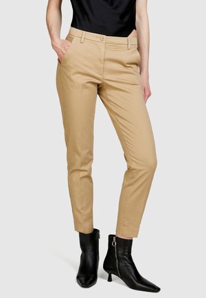 Nainen pukeutuneena beigenvärisiin slim fit -housuihin, joissa on etutaskut, ja mustiin nilkkureihin, joissa on sivuvetoketjut, seisomassa yksivärisen taustan edessä.