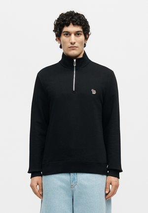 Młody mężczyzna z kręconymi włosami nosi czarny pulower z suwakiem do ćwiartki i logo zebry oraz jasnoniebieskie dżinsy, stojąc na białym, jednolitym tle.