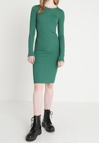 Grön långärmad figursydd klänning i elastiskt material, med rund halsringning och knälång nederkant, tillsammans med svarta snörade platåstövlar.
