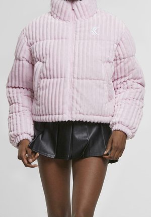 Kobieta w jasno różowej, teksturowanej kurtce puchowej z zamkiem błyskawicznym, ubrana w czarną plisowaną mini spódnicę ze skóry, stojąca na jednolitym tle.