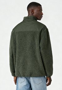 Giacca in pile verde con corpo soffice e testurizzato e parte superiore in tessuto liscio. Colletto alto e vestibilità comoda, vista da dietro.