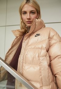 Rožinė pūkinė striukė su blizgiu paviršiumi, aukšta apykakle ir Nike logotipu kairiajame krūtinės pusėje. Modelis dėvi tamsų megztinį po ja.