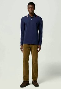 Camisa polo de manga longa azul-marinho com colarinho contrastante vermelho e branco, combinada com calças chino castanhas e sapatos escuros, de pé contra um fundo neutro.