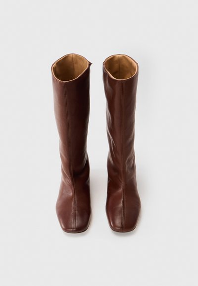 Bottes en cuir marron montantes avec une texture lisse, bouts carrés et coutures minimales. Intérieur doublé d'une couleur beige clair.