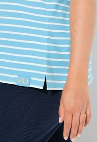 Modrý pruhovaný bavlněný tričko s postranním rozparkem, se malou žlutou nášivkou na spodním lemu, doplněné tmavými ribkovanými kalhotami.