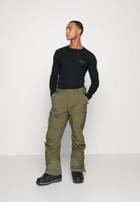 Camisa negra de manga larga y pantalones cargo verdes olivo con bolsillos, combinados con botas de nieve negras, sobre un fondo neutro.