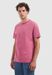 Hellrosa Baumwoll-T-Shirt mit kurzen Ärmeln und rundem Halsausschnitt. Verfügt über ein kleines bedrucktes Logo "PURE PATH" nahe dem Saum. Schlichtes Design.