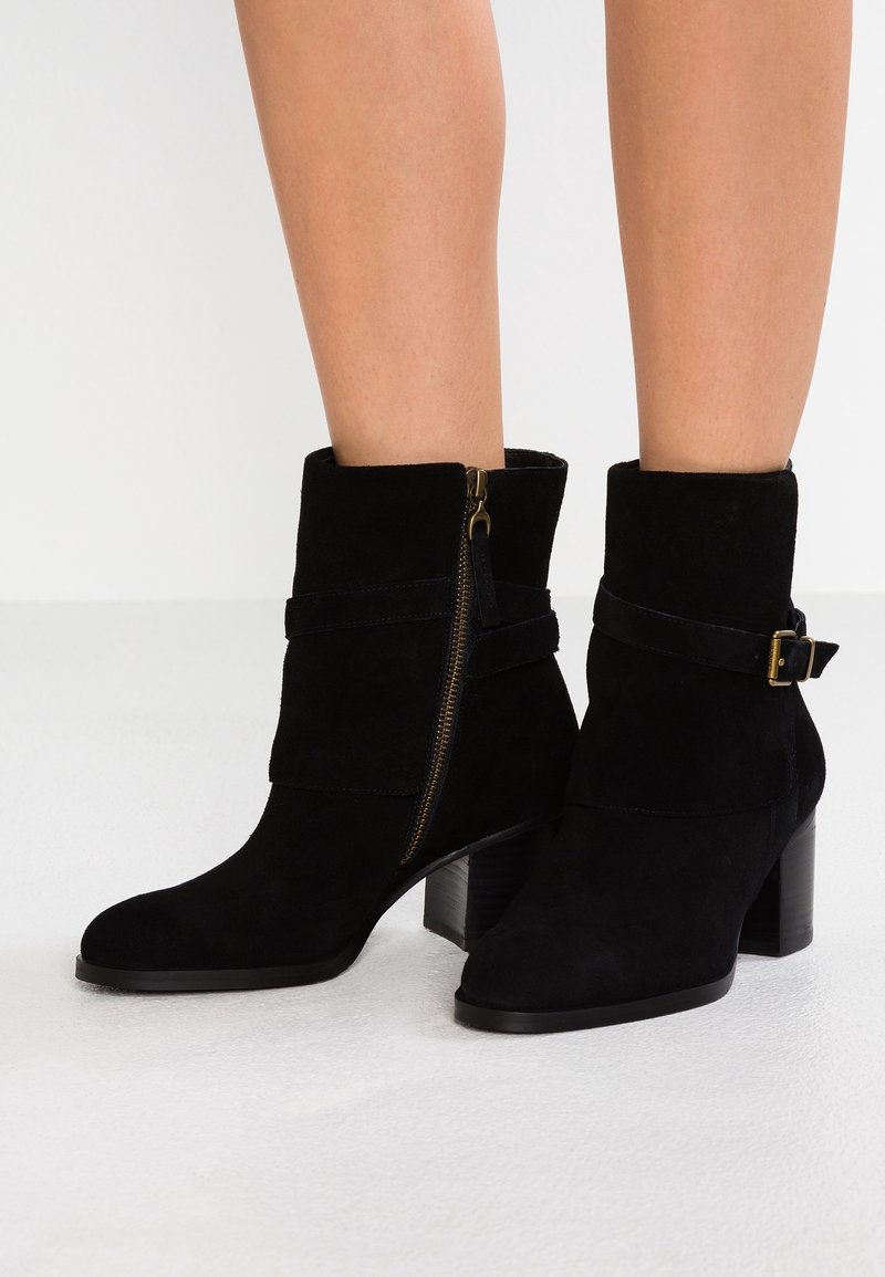 Botines de ante negros con tacón bloque, que cuentan con una cremallera lateral y una correa con detalle de hebilla. Textura suave y diseño ajustado.
