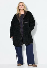 Cappotto in pelliccia sintetica nera con ampio colletto, rifinito con bordi marroni, bottoni frontali e due tasche laterali. Indossato sopra un top a righe blu navy e pantaloni.
