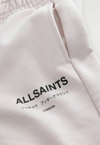 Zbliżenie na jasnoróżowy materiał z czarnym napisem "ALLSAINTS", znakami japońskimi i napisem "LONDON" nadrukowanym na szwie kieszeni.