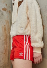 Fuzzy krämfärgad pullover med en texturerad yta och huva, i kombination med röda shorts med vita sidOSTRECK och en Adidas-logotyp.