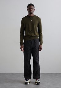 Olivenfarget strikkegenser med et strukturert diagonalmønster, parret med svarte vide bukser og multicolor sneakers.