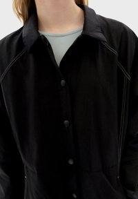 Czarna kurtka zapinana na guziki z kołnierzowym dekoltem, z białym, kontrastującym przeszyciem, teksturowaną tkaniną i dwoma guzikami z przodu.