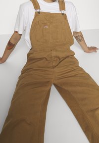 Personne assise les jambes étendues, portant une salopette marron et un t-shirt blanc, montrant des tatouages colorés sur les deux avant-bras.