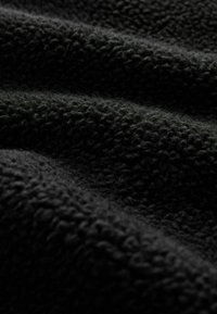 Weicher, schwarzer Fleece-Stoff mit dichter, lockiger Textur und sanften Falten, die Schatten und Lichtreflexe erzeugen.