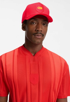 Man draagt een rood gestreept shirt en een rode honkbalpet met een Spaans vlagembleem op een witte achtergrond.