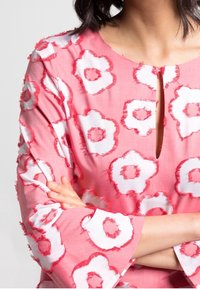 Blouse florale rose avec des motifs de fleurs blanches, dotée d'un col en forme de goutte d'eau et de détails texturés effilochés le long des manches.