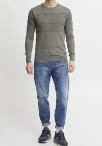 Uomo che indossa un maglione grigio a trama, jeans blu risvoltati, calze bianche e sneakers blu navy, in piedi su uno sfondo bianco semplice.