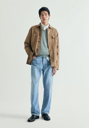 Mladý muž stojící, oblečený v hnědém kabátě, šedém svetru přes bílou košili, světle modrých džínách a černých botách, na jednoduchém světlém pozadí.