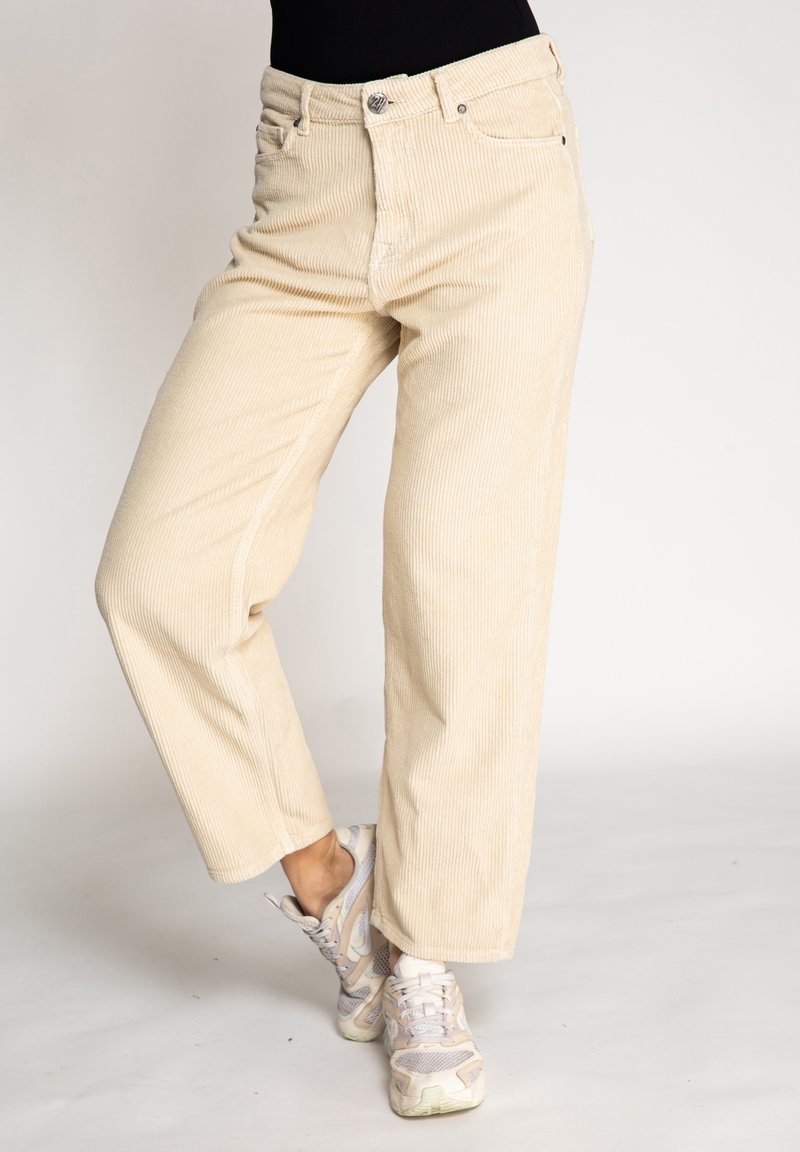 Beige corduroy wide-leg pants with a textured surface, front pockets, and a metal button closure, paired with light-colored sneakers.