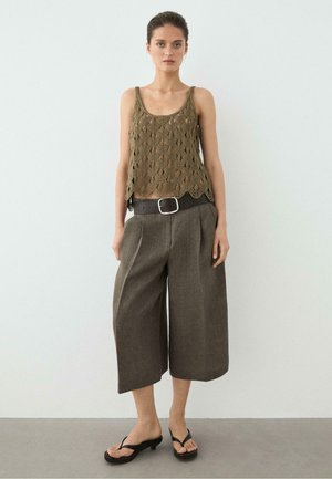 Junge Frau steht vor einer weißen Wand, trägt ein olivgrünes gehäkeltes Top, braune weit geschnittene kurze Hosen mit schwarzem Gürtel und schwarze Sandalen mit Absatz.