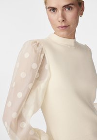 Pull en tricot couleur crème doux avec une texture côtelée et un col montant, présentant des manches bouffantes transparentes ornées de pois blancs.
