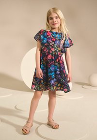 Lilleline kleit lühikeste volangidega varrukatega; tumedat tausta ilmestavad roosad, sinised ja kollased värvilised lilled. Sobitatud heledate, rihmikandadega sandaalidega.