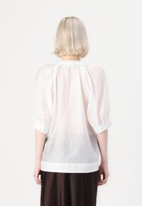 Blusa bianca leggera e semitrasparente con maniche a sbuffo, dettagli in crochet sulla schiena e orlo dritto. Texture liscia.