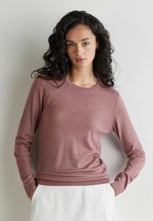 Kvinde med krøllet mørkt hår iført en langærmet støvet rosa sweater og hvide bukser står op ad en hvid væg.