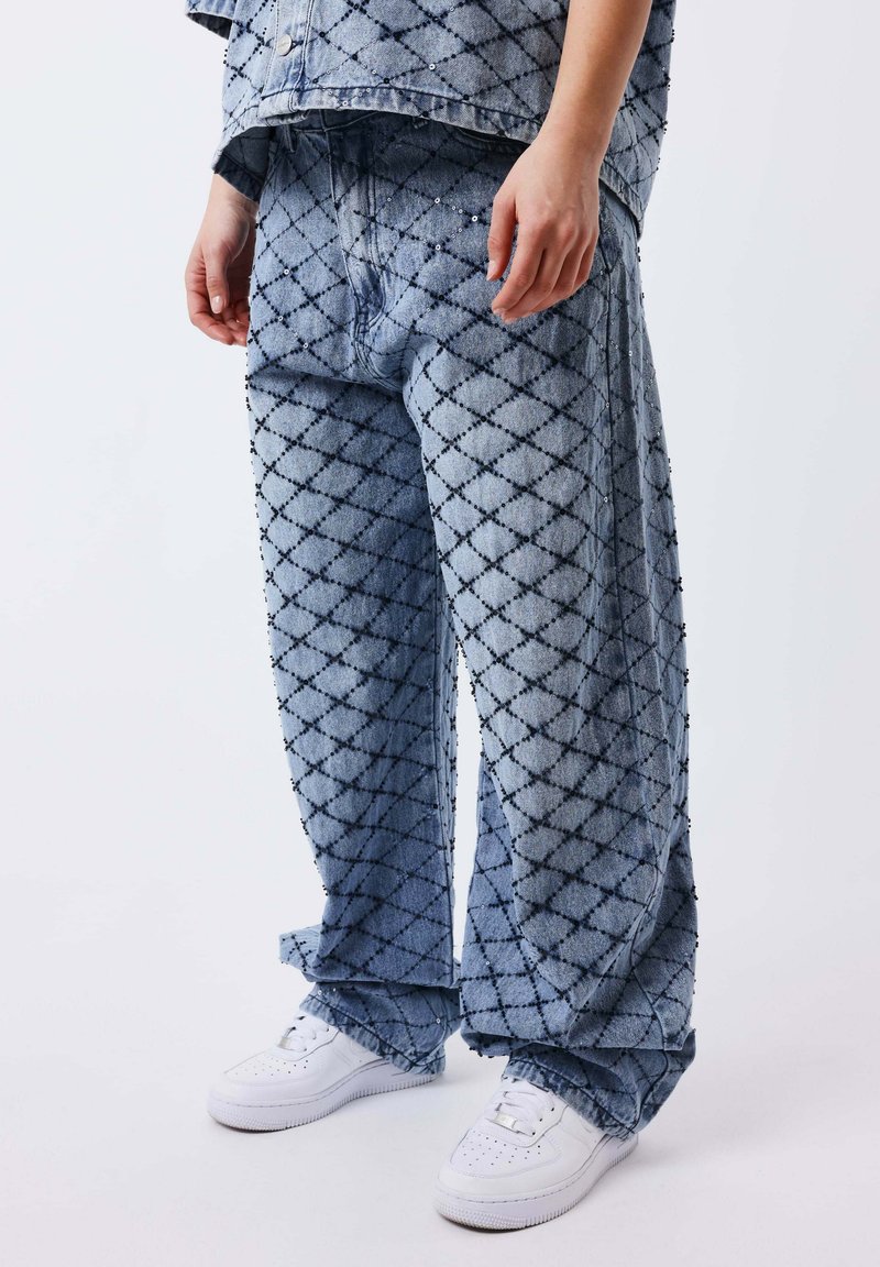 Personne portant un pantalon bleu ample et une veste assortie avec un motif de losanges noirs, associée à des baskets blanches sur fond blanc.