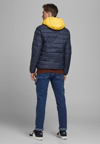 Navy quilted jacket with a yellow hood, paired with brown top and blue jeans. The jacket features a sleek, puffy design.