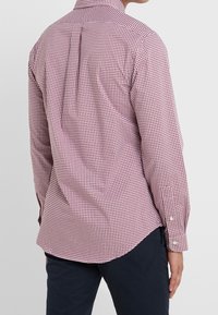 Man i en långärmad skjorta i vinröd och vit rutigt mönster med mörka byxor, sedd från ryggen mot en enkel bakgrund.