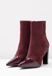 Bottes courtes bordeaux en daim avec un bout pointu en cuir verni. Elles présentent un talon carré et une texture lisse.
