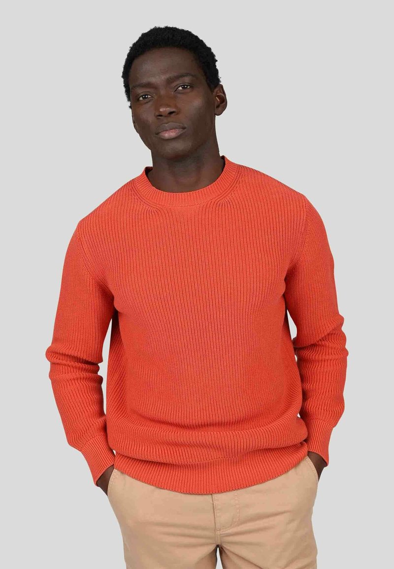 Pull en tricot côtelé orange avec un col rond et des manches longues, présentant une finition texturée et une coupe décontractée. Un pantalon beige est visible.