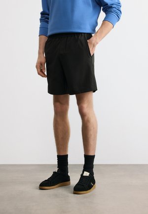 Άνδρας που φοράει μαύρο σορτς με ελαστική μέση, μπλε πουκάμισο με μακριά μανίκια, μαύρες κάλτσες και μαύρα αθλητικά παπούτσια με καουτσούκ σόλες, στέκεται σε γκρι πάτωμα.