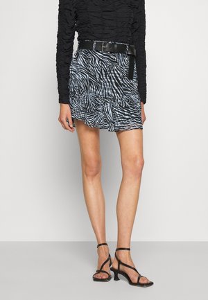 Femme portant un haut noir à manches longues froncées, une jupe courte à imprimé zèbre avec ceinture noire, et des sandales à talons noires à fines lanières.