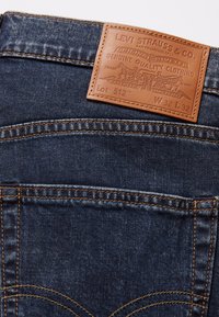 Jeans de mezclilla oscura con un parche de cuero marrón etiquetado "LEVI STRAUSS & CO." Presentan costuras amarillas y bolsillos estándar. Modelo 512, tamaño 32x32.
