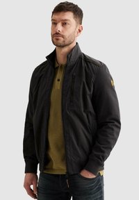 Homme aux cheveux courts et foncés avec une barbe, portant une veste noire zippée sur un polo vert olive, debout avec une main dans la poche.