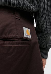 Pantalones de algodón marrón con un bolsillo trasero que tiene una etiqueta blanca y un logo naranja. Suéter gris de punto texturizado parcialmente visible en la parte superior.