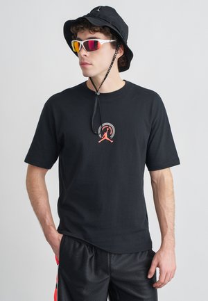 Črna bombažna majica s rdečim grafičnim logotipom na prsih, kombinirana s črnimi športnimi kratkimi hlačami in črnim klobučkom. Videz dopolnjujejo zrcalna sončna očala.