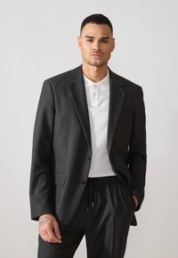 Blazer entallado gris oscuro con dos botones, solapas con muesca y bolsillos laterales, combinado con una camiseta polo blanca y pantalones oscuros a juego.
